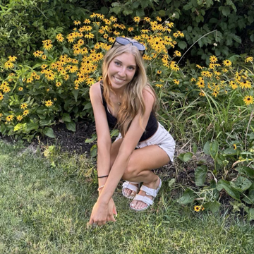 Young caucasian woman bent down next to yellow flowers in white shorts, black tank top, and white sandals. Blonde long hair. 
