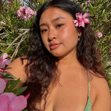 Young person of color with dark brown wavy hair and a pink flower in it. 
