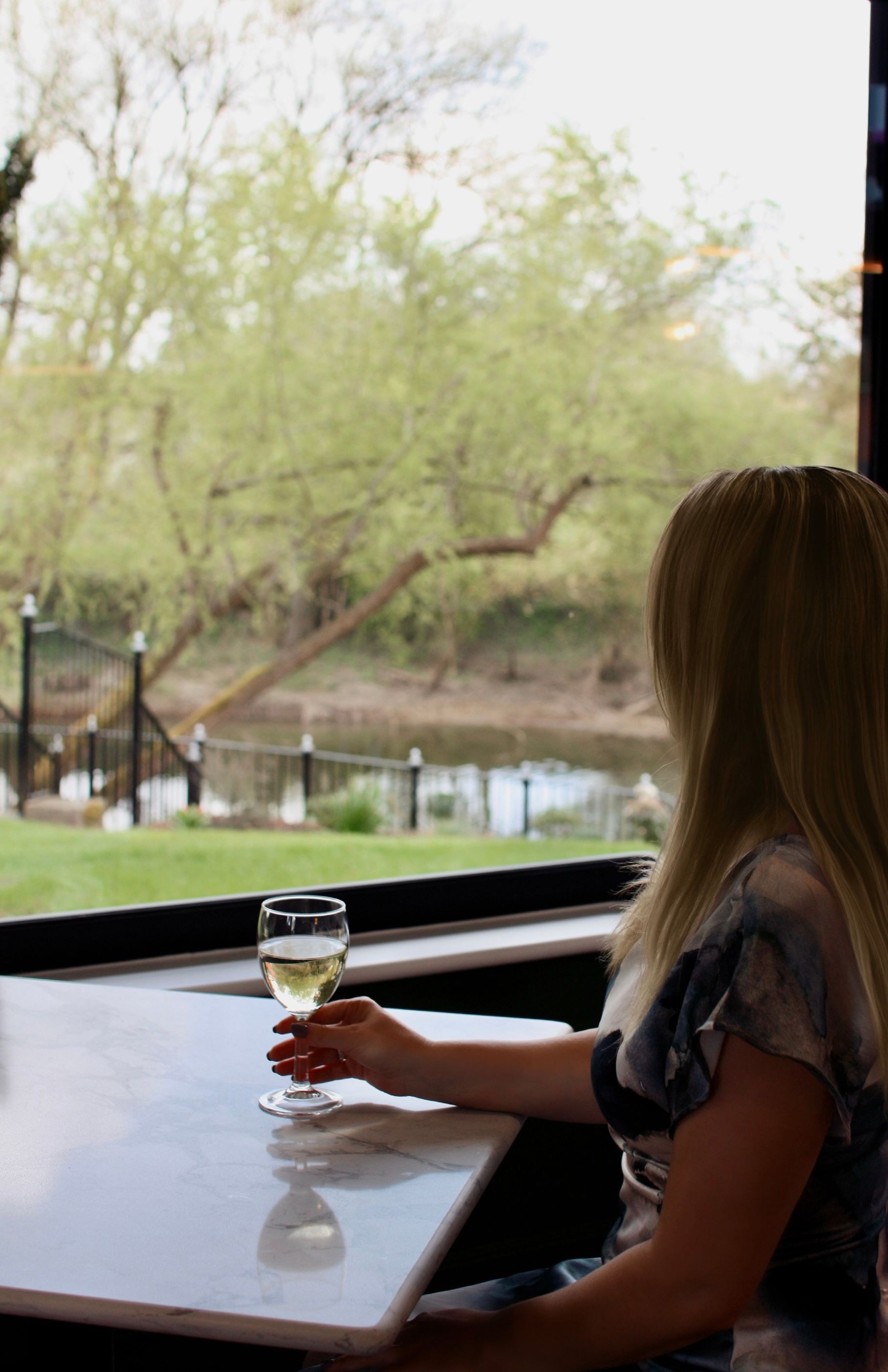 Woman sitting at a table by a window, holding a glass of white wine, looking out at a river.