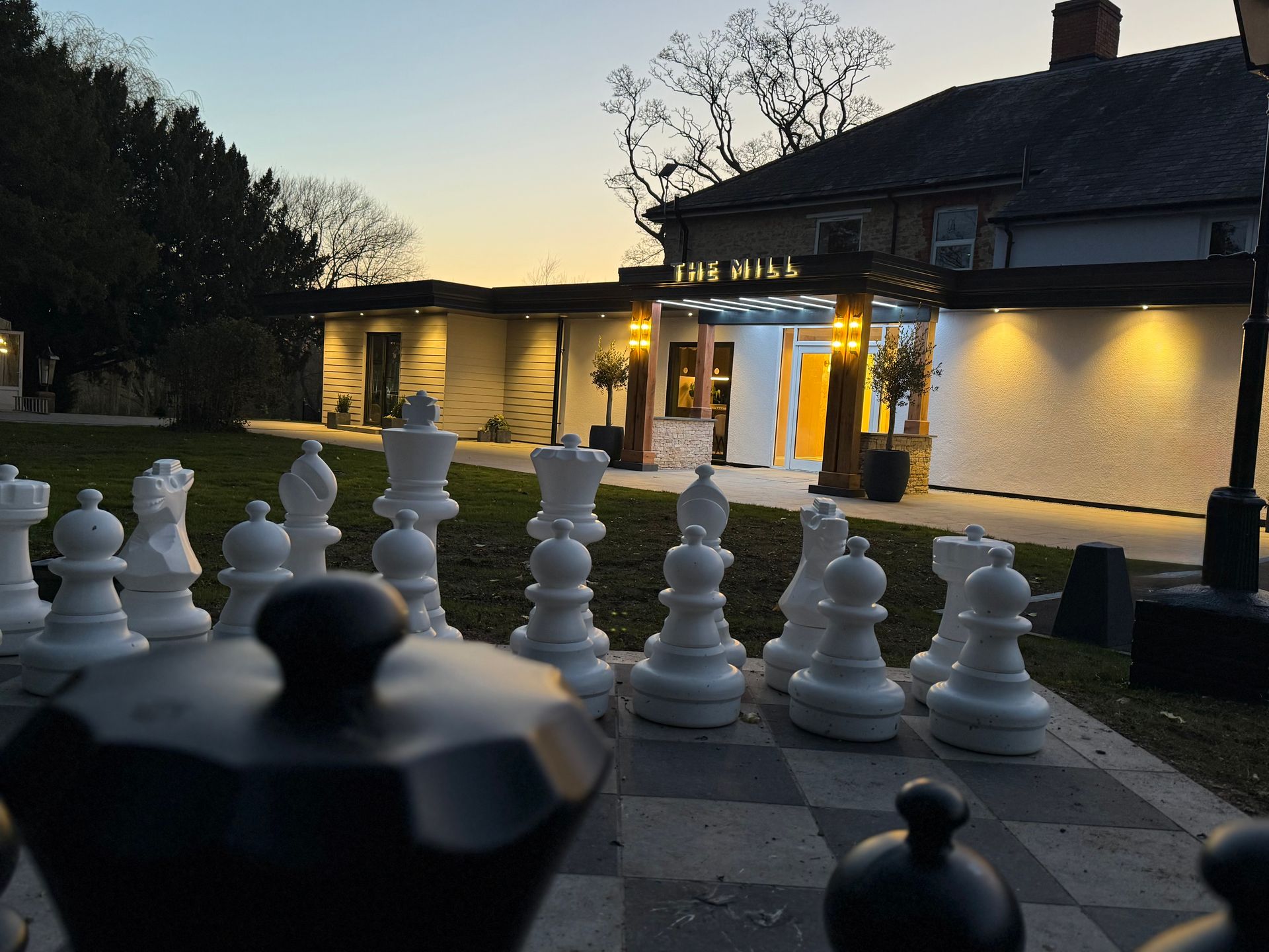 Large outdoor chess set with a building in the background, illuminated at dusk.