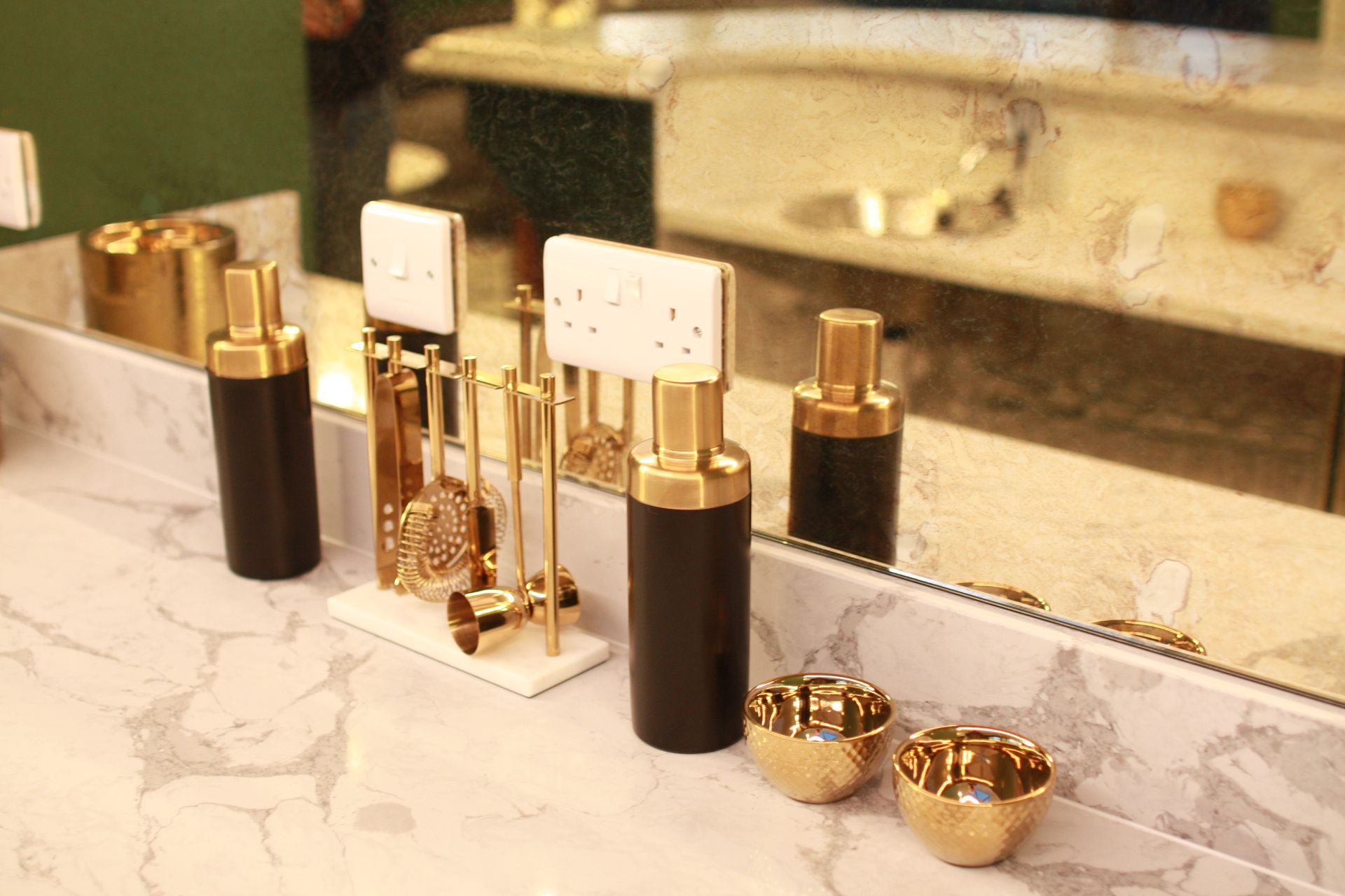 Bar setup with gold and black bar tools on a marble counter, reflected in a mirror.