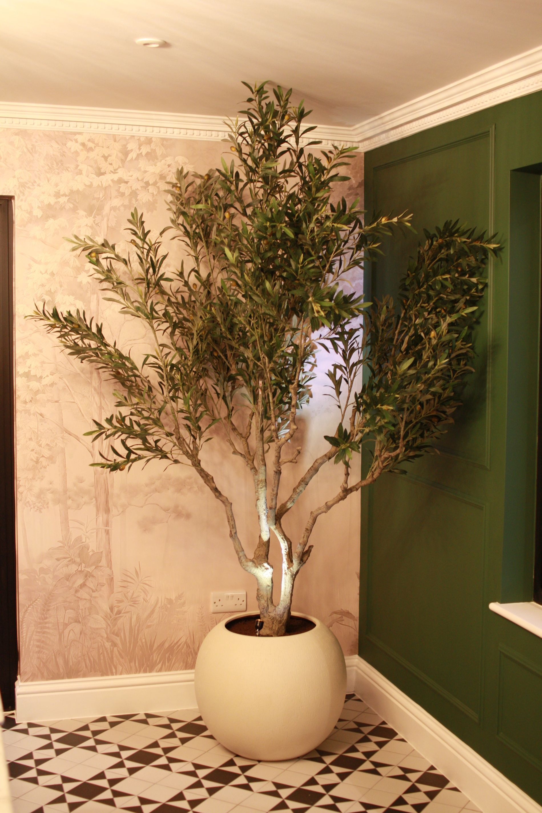 Large artificial olive tree in a white spherical planter against patterned floor and green panelled wall.