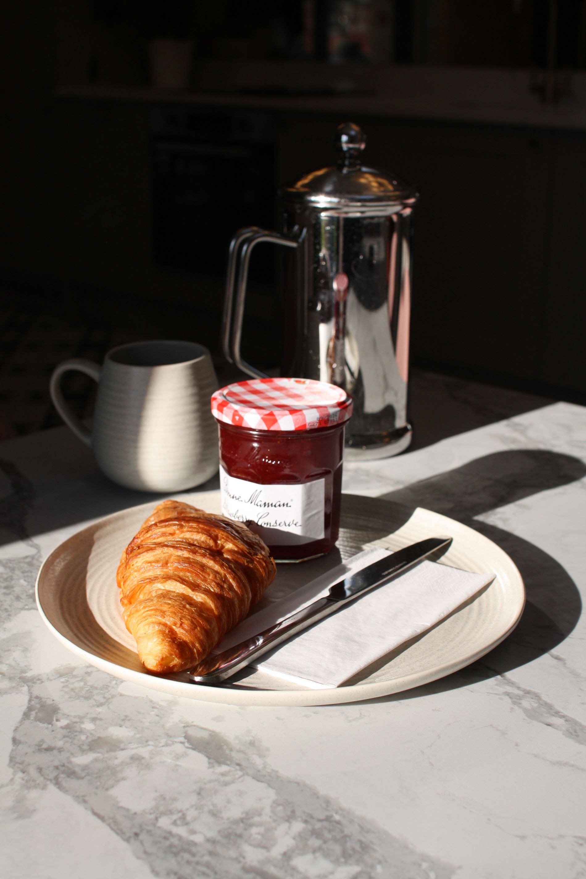 Croissant, jam, knife, and mug on a plate with coffee press. Sunny marble tabletop.