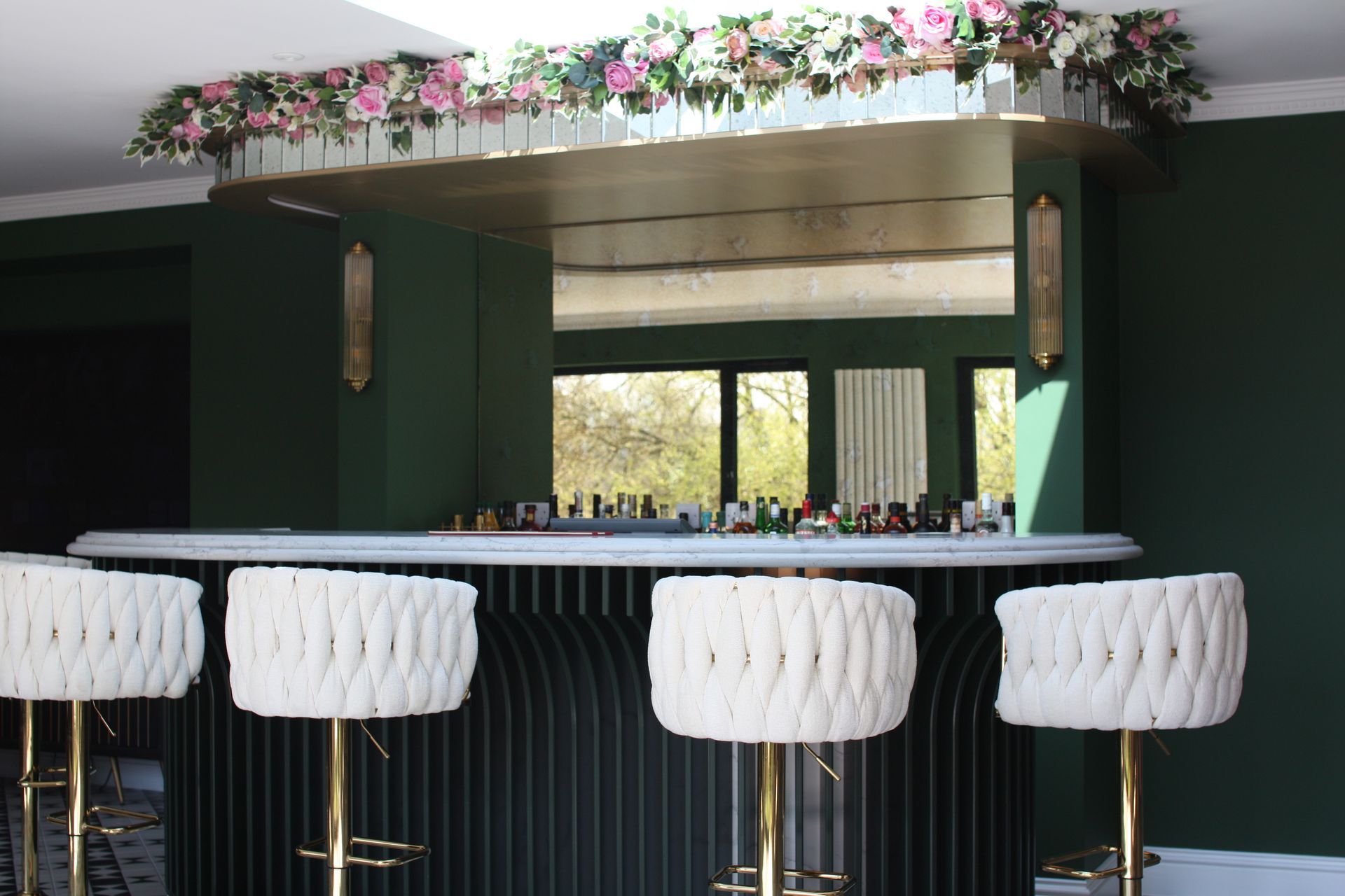 Green bar with white stools, gold accents, floral decor, and a mirror.