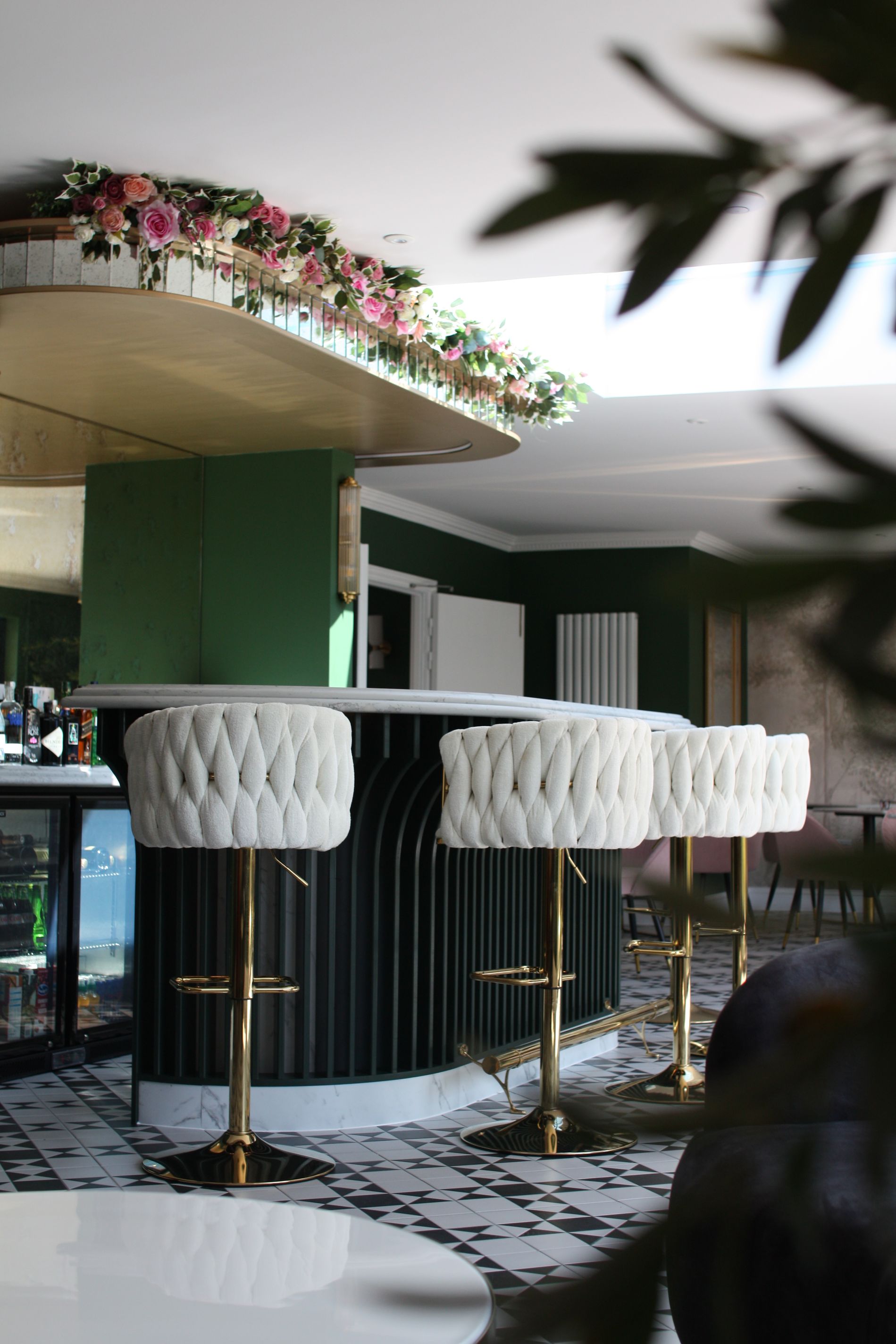Bar with white upholstered stools, black bar, gold accents, floral garland, and green wall.