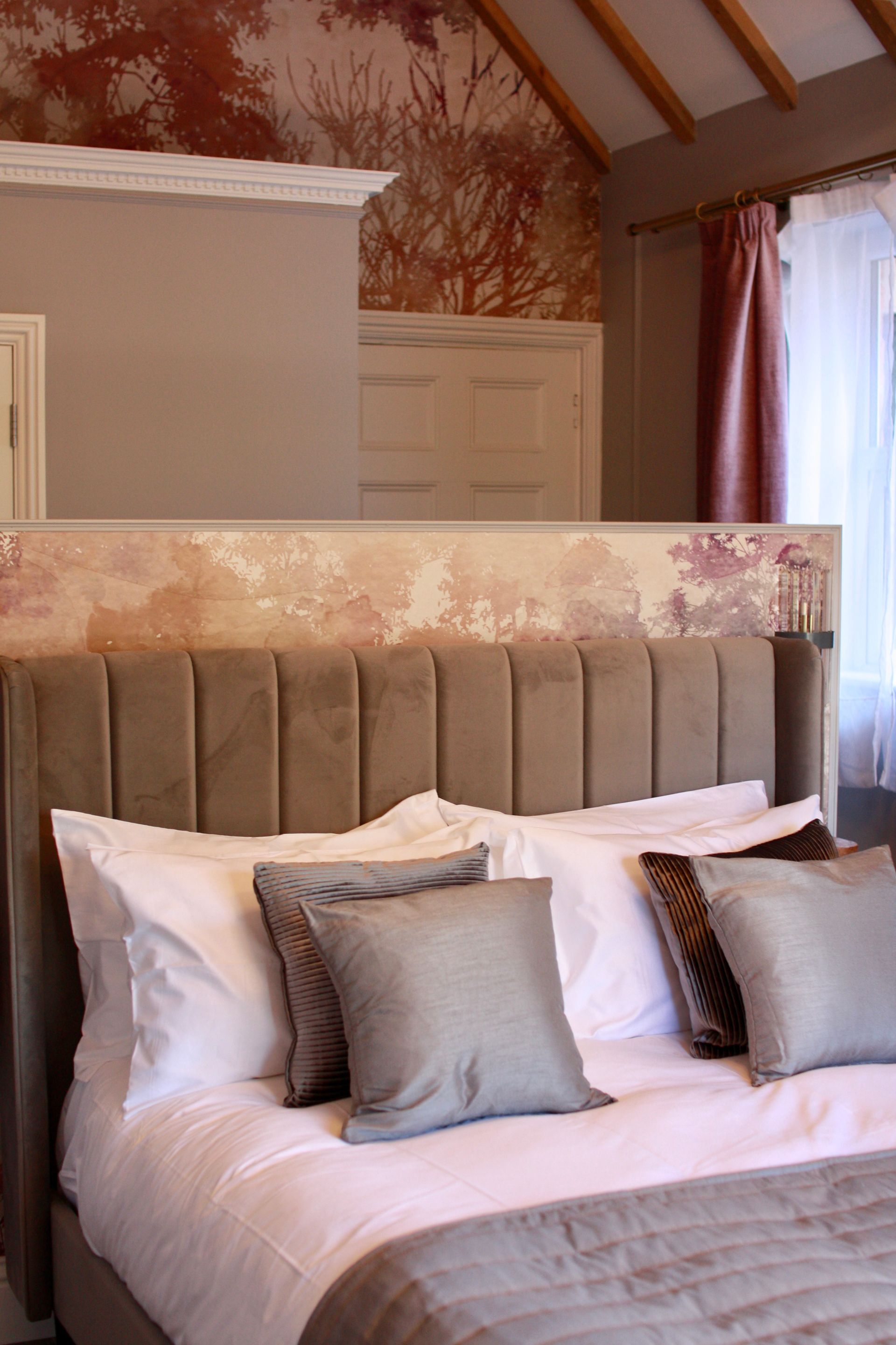 Cozy bedroom with upholstered bed, white linens, and decorative pillows. Soft, warm colors; floral wallpaper.