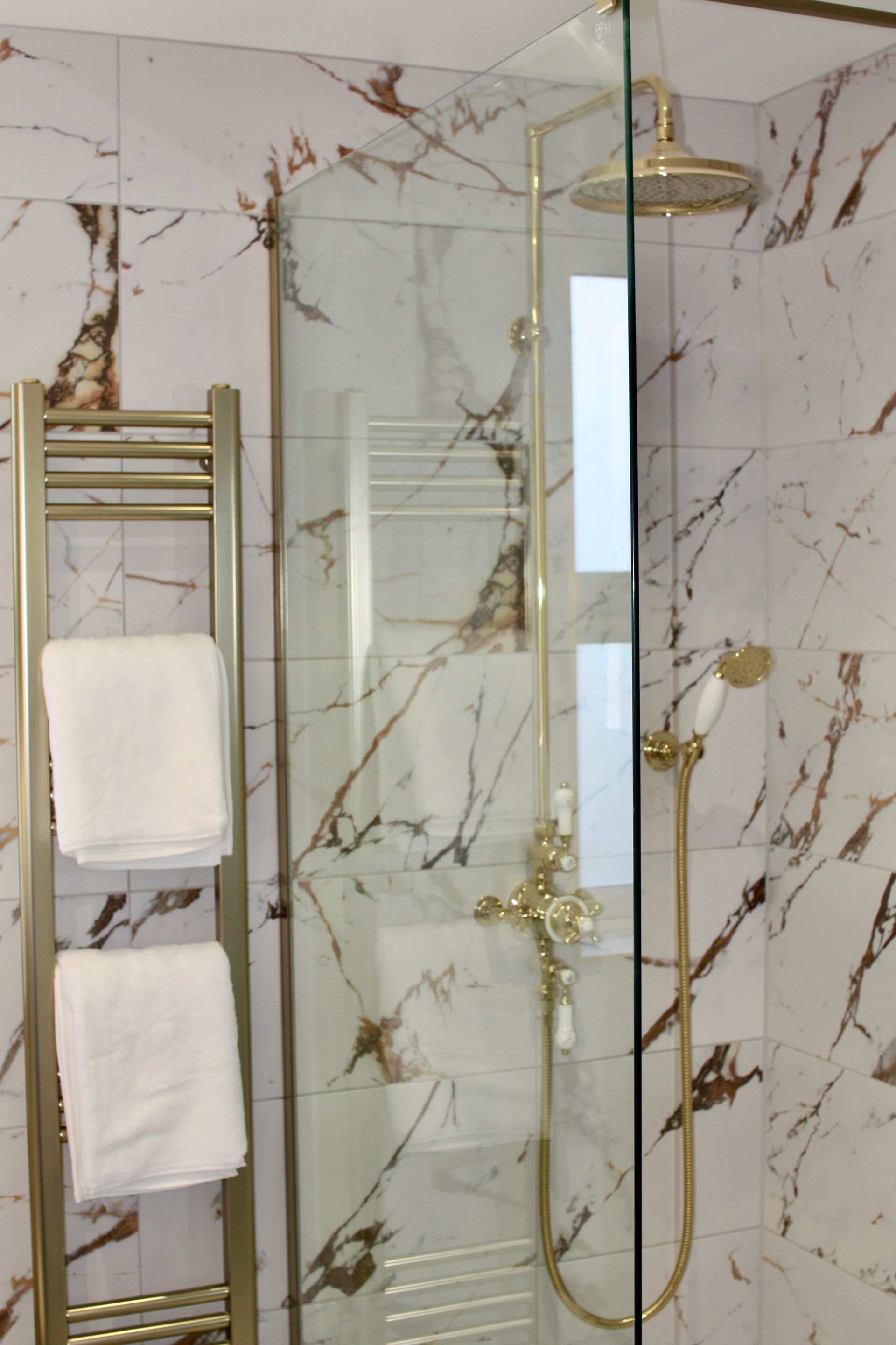 Gold-toned shower fixture and towel rack in a bathroom with marble-look tile. Two white towels hang on the rack.
