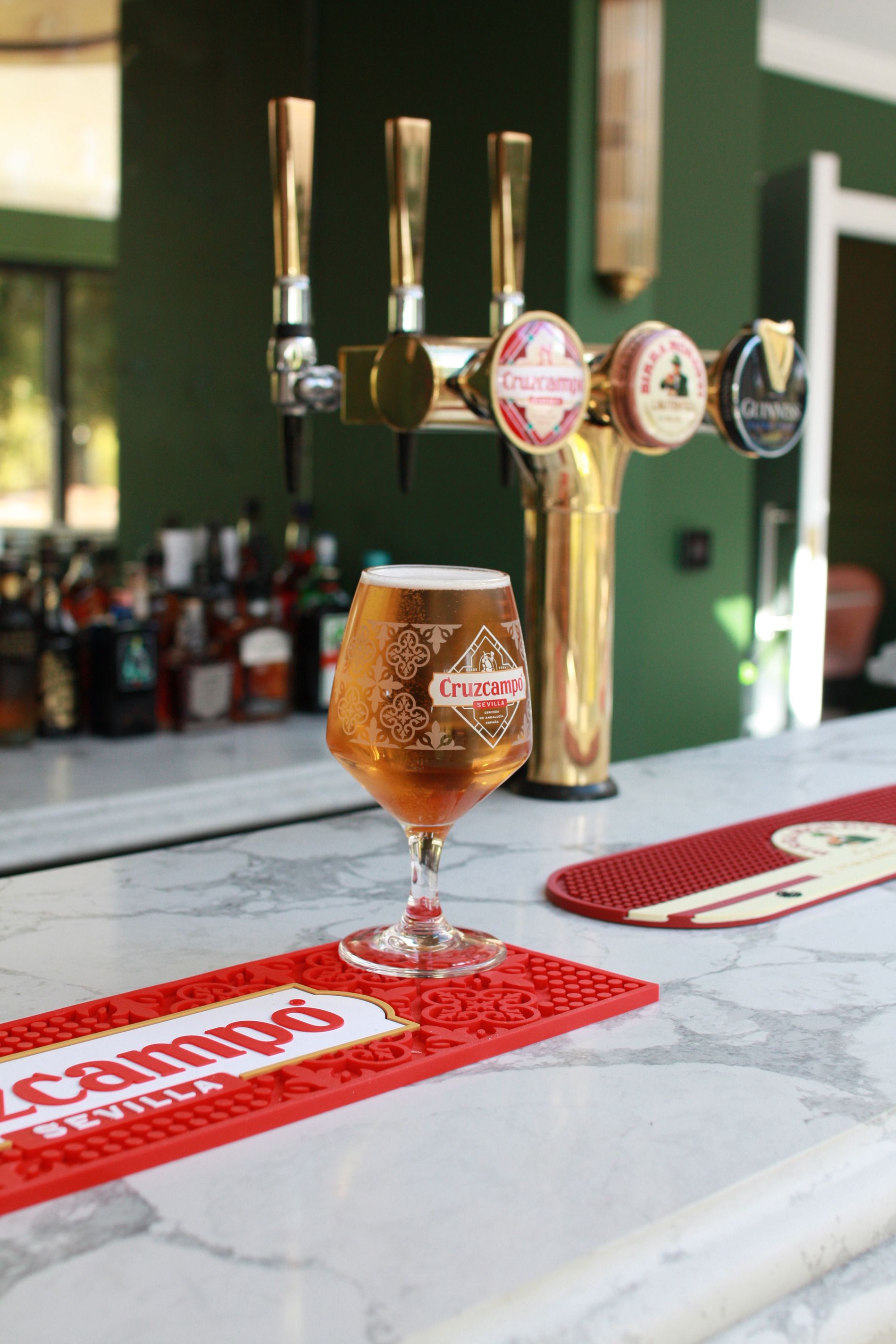 A beer glass filled with amber beer sits on a bar in front of golden beer taps.