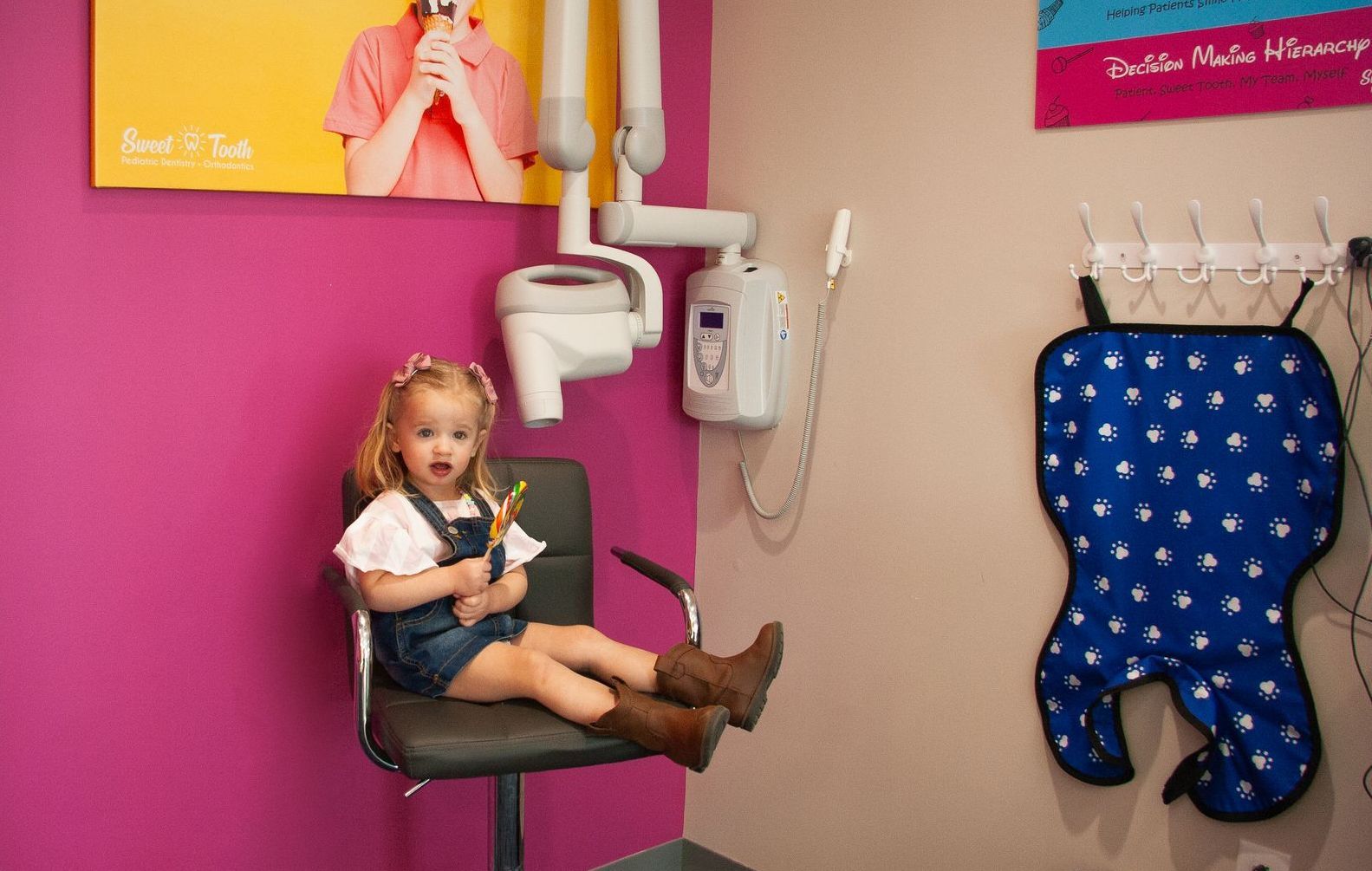 A little girl sitting on a chair in a dental office holding a lollipop. 