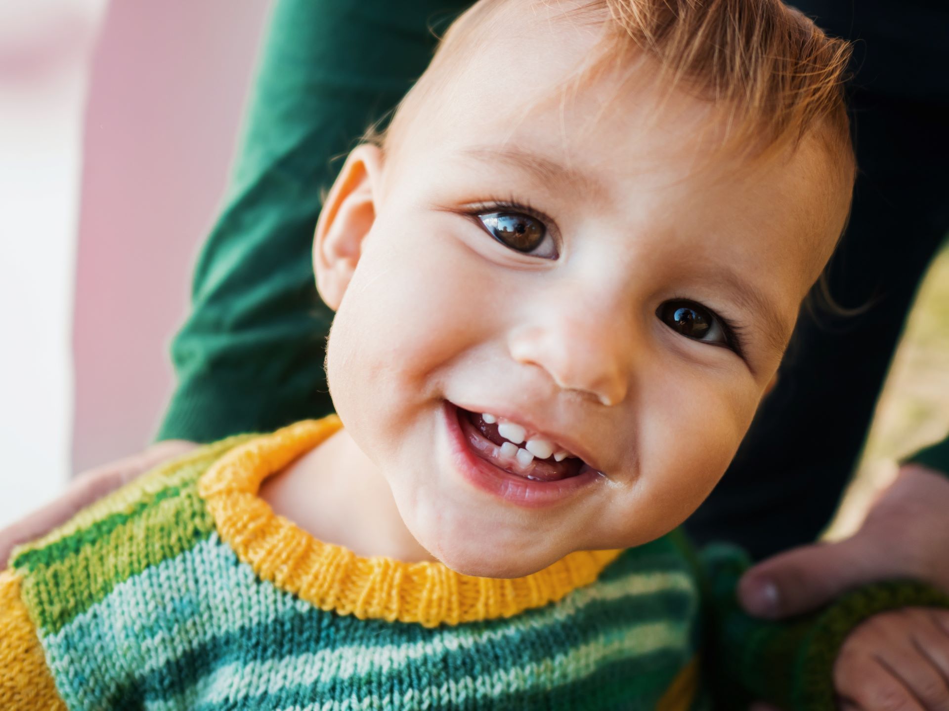 Smiling baby with brown eyes wearing a green and yellow sweater; held by a person.