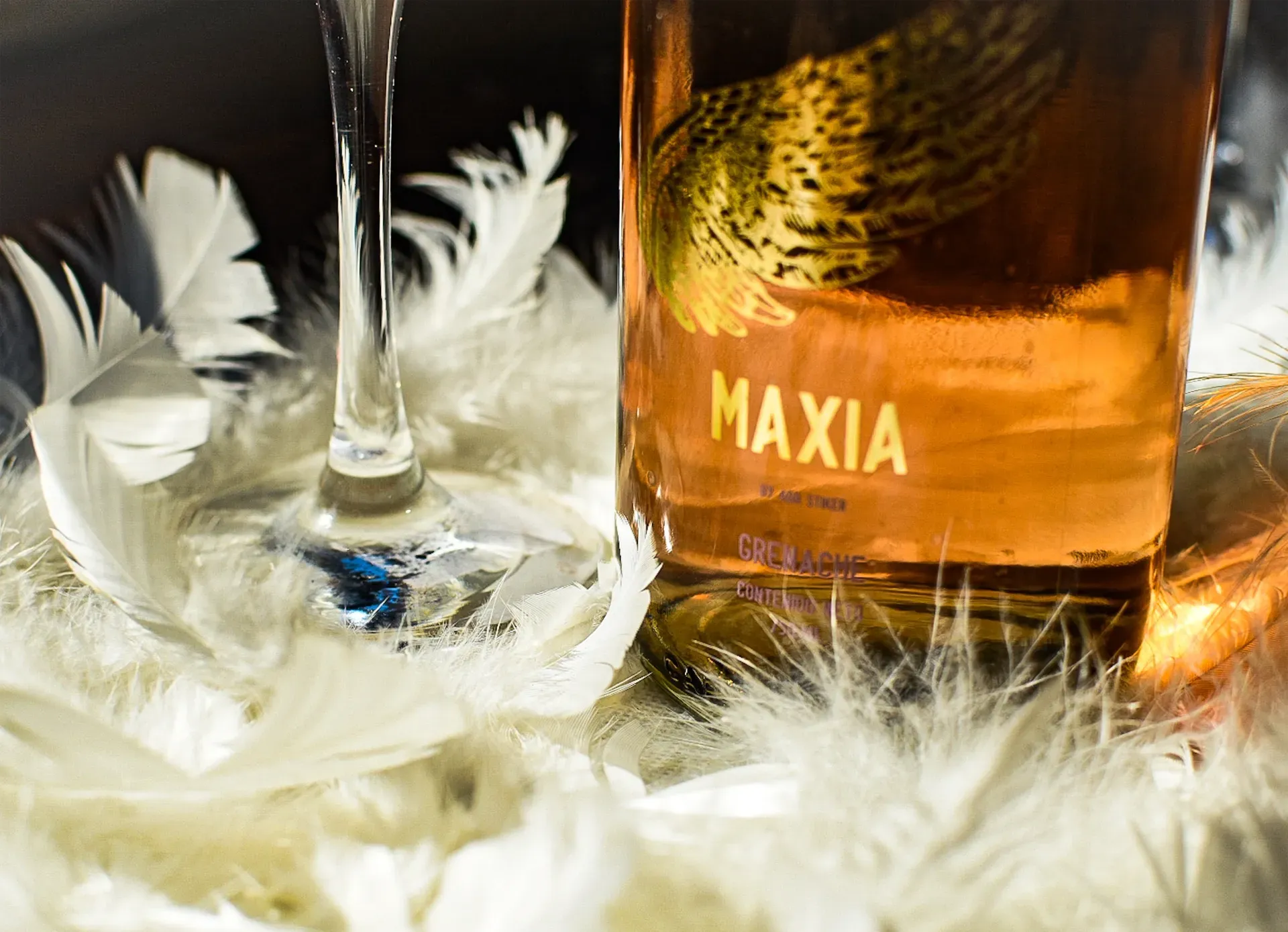Botella de la bebida “MAXIA” al lado de una copa de vino, apoyada sobre plumas blancas; el tallo de la copa está en foco.