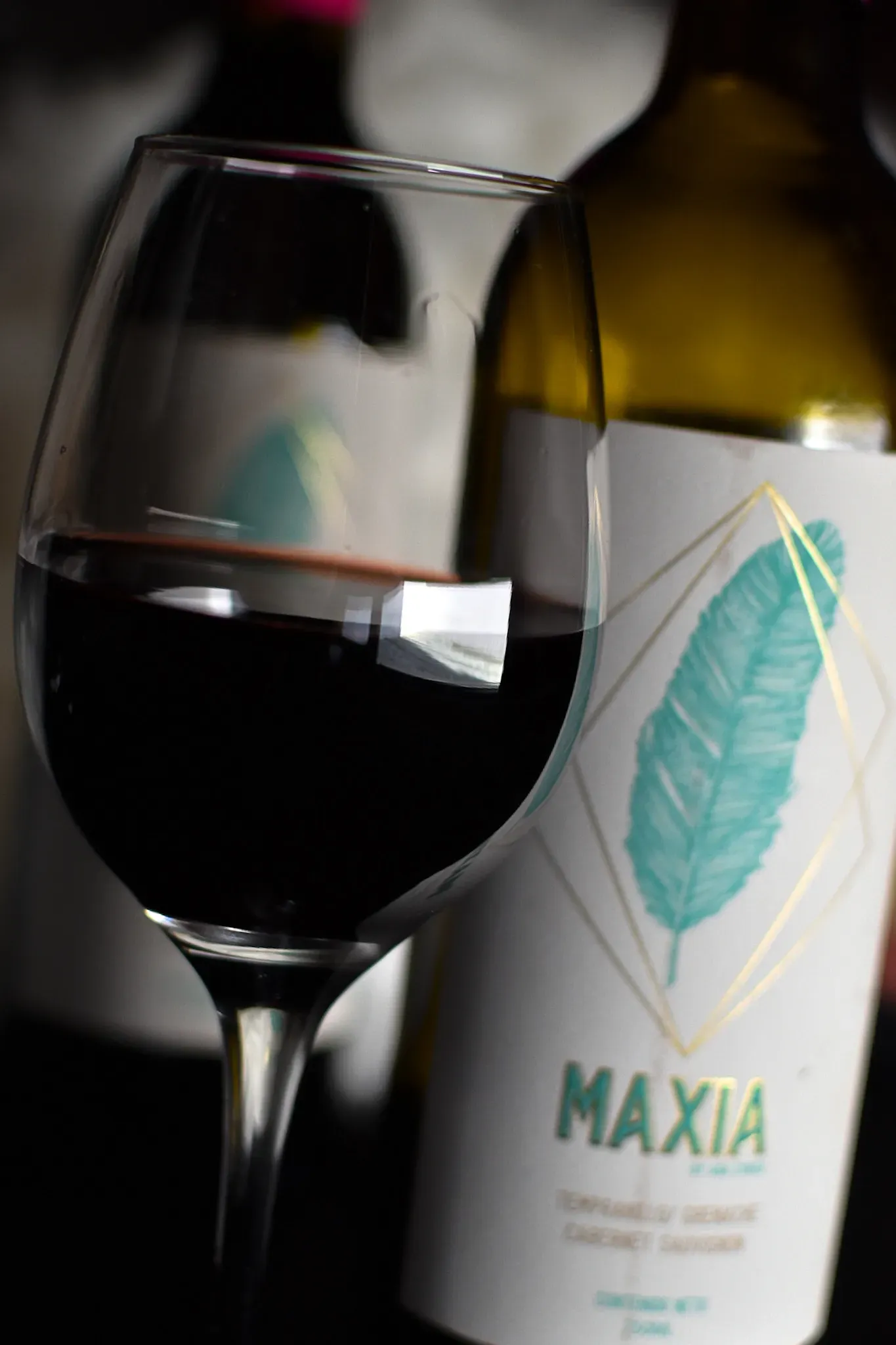 Copa de vino con vino tinto, botella con diseño de hoja turquesa, etiqueta “MAXIA”.