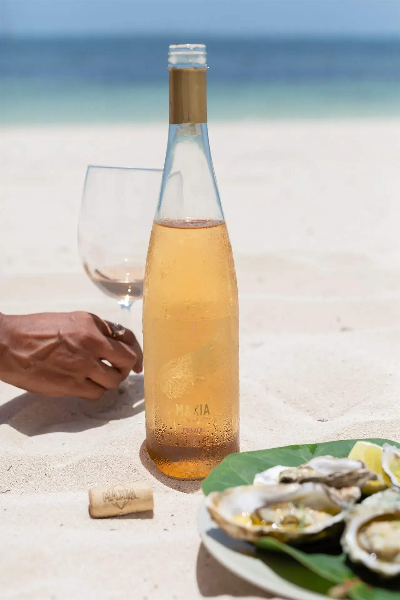 Una botella de rosado maxia y ostras en un plato en una playa de arena. Una mano toma una copa de vino.