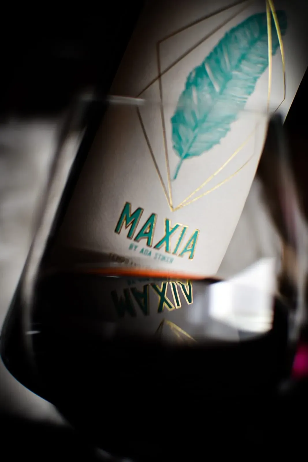 Botella de vino MAXIA en copa; la etiqueta presenta un diseño de plumas verde azulado y formas geométricas doradas.