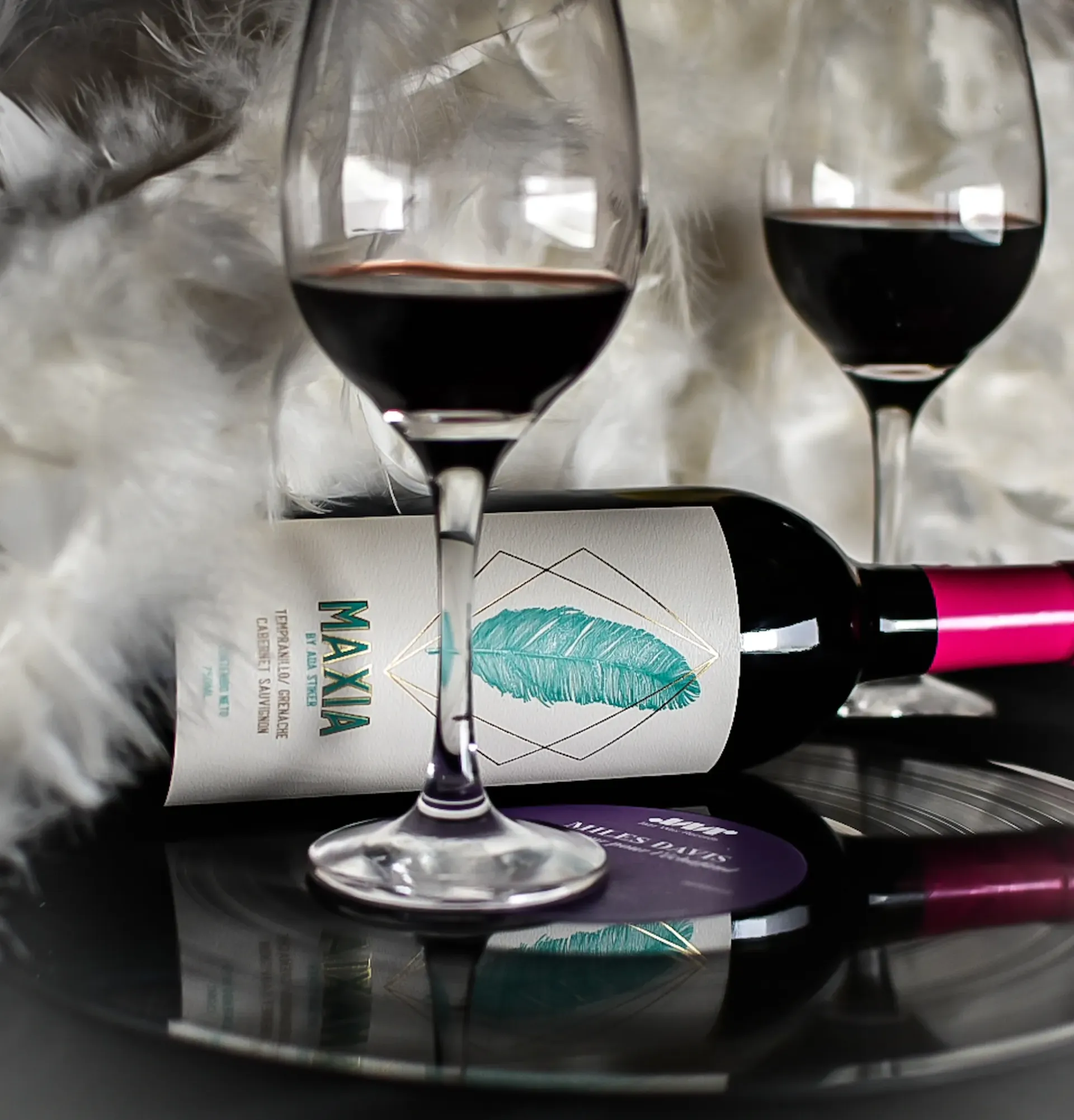 Dos copas de vino y una botella de vino tinto maxia, con un fondo de plumas.