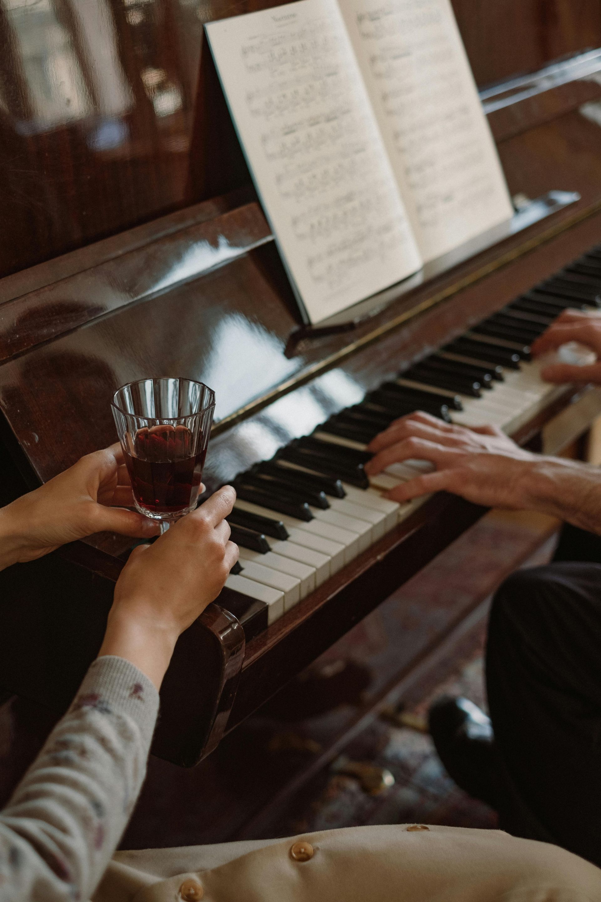 Las manos tocan las teclas del piano; una sostiene una copa de vino maxia cerca de una partitura.