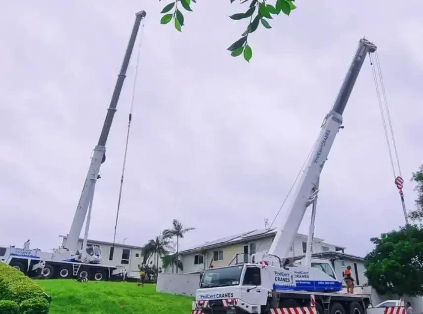 Two Crane Trucks Are Sitting On Top Of A Grassy Hill In Front Of A Building — ProCert Cranes In Sunshine Coast, QLD