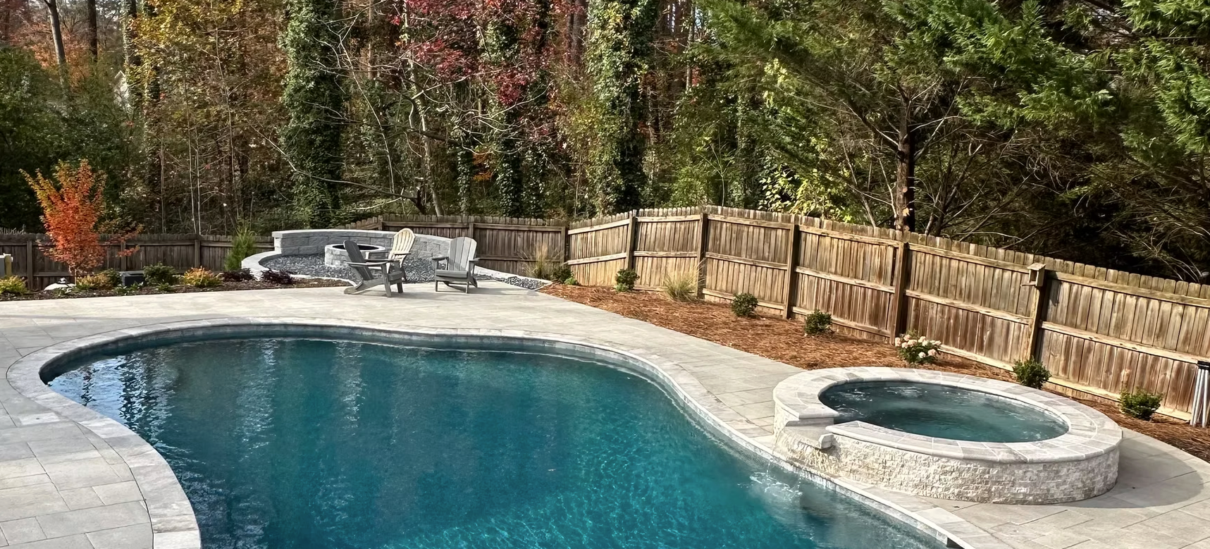 A large swimming pool with a hot tub in the backyard surrounded by trees.