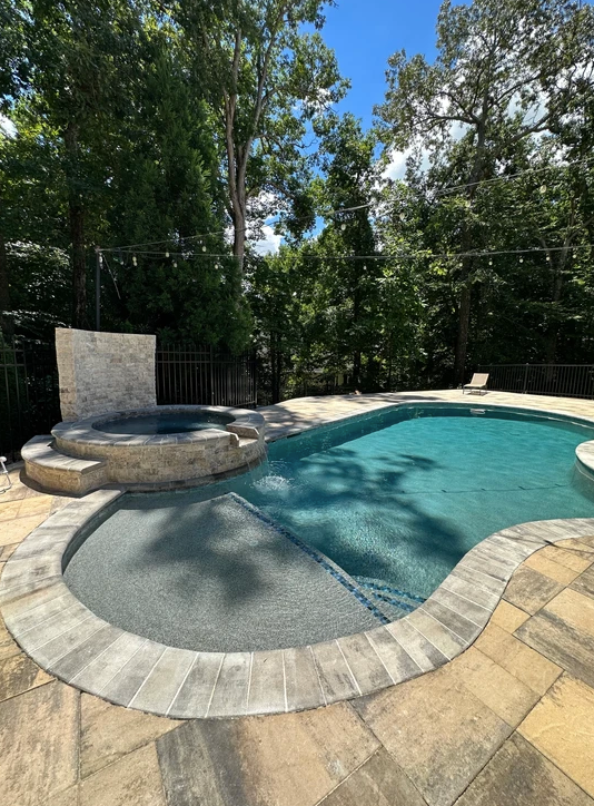 A large swimming pool with a hot tub in the backyard surrounded by trees.