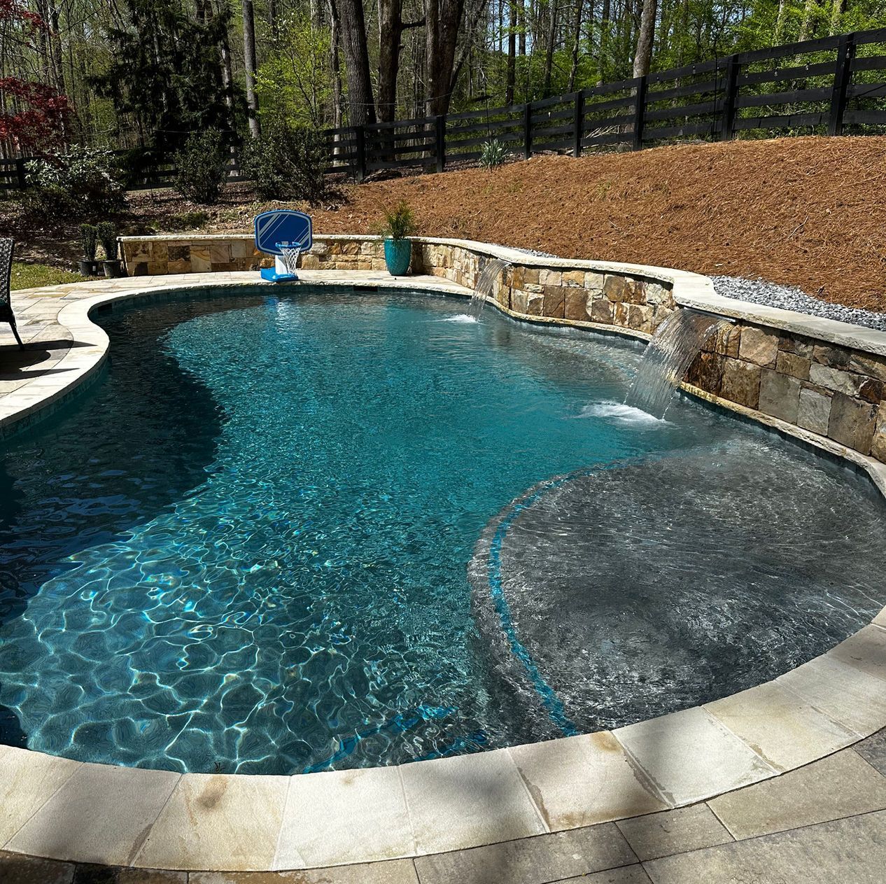 A large swimming pool is surrounded by a stone wall and a fence.