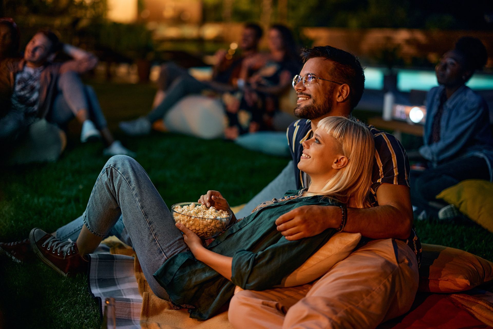 A man and a woman are sitting on the grass eating popcorn and watching a movie.