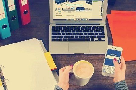 A person is holding a cup of coffee in front of a laptop and a cell phone.