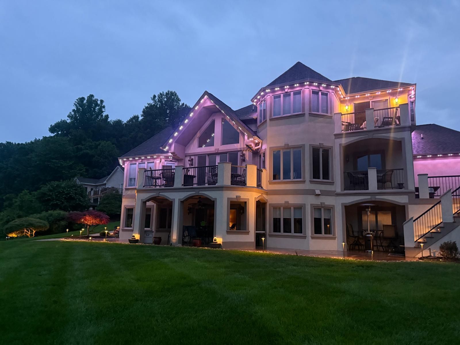 Large house lit with pink and white lights at dusk, green lawn, trees in background.