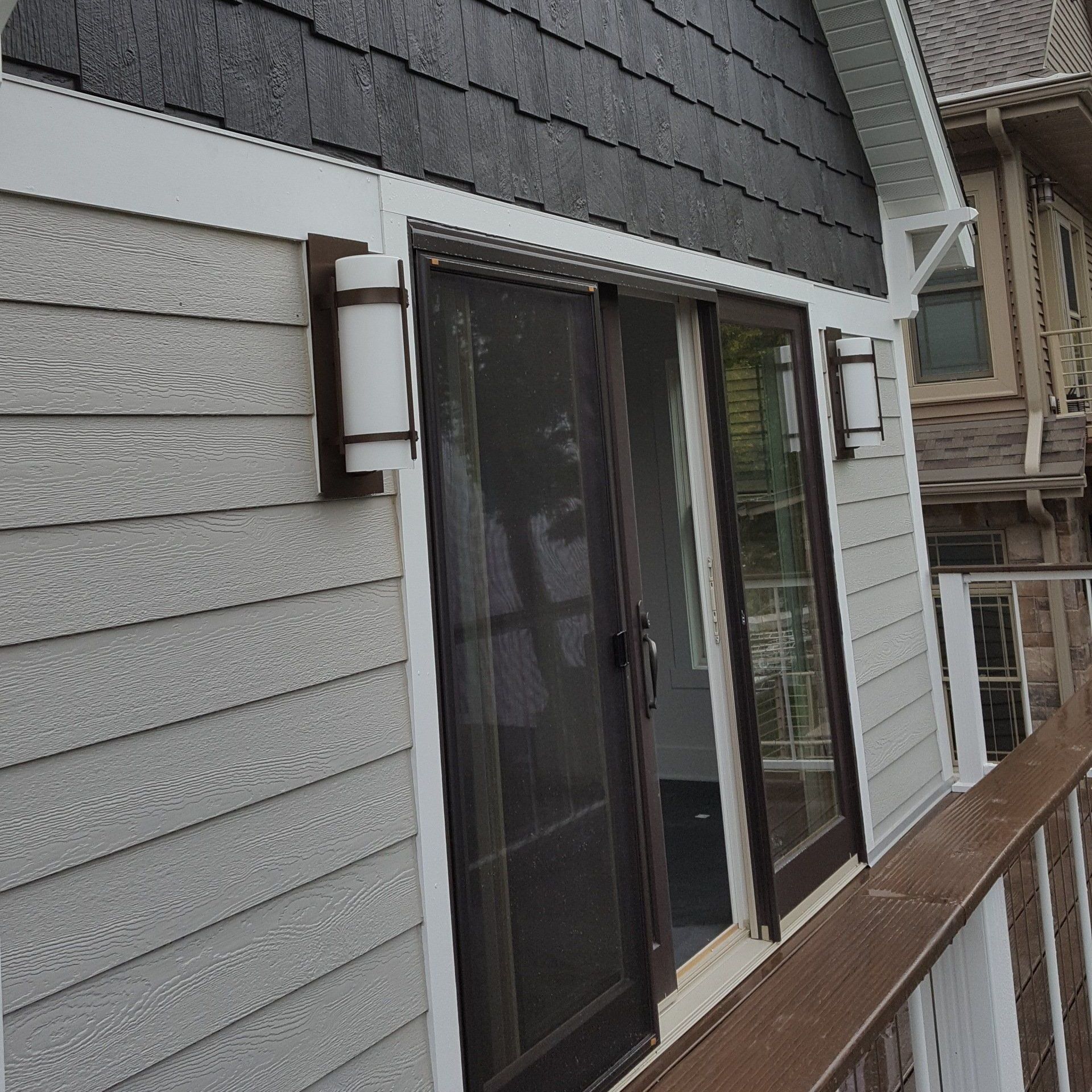 Exterior of house with sliding glass door and two wall-mounted lights; gray siding, dark trim, and dark roof.