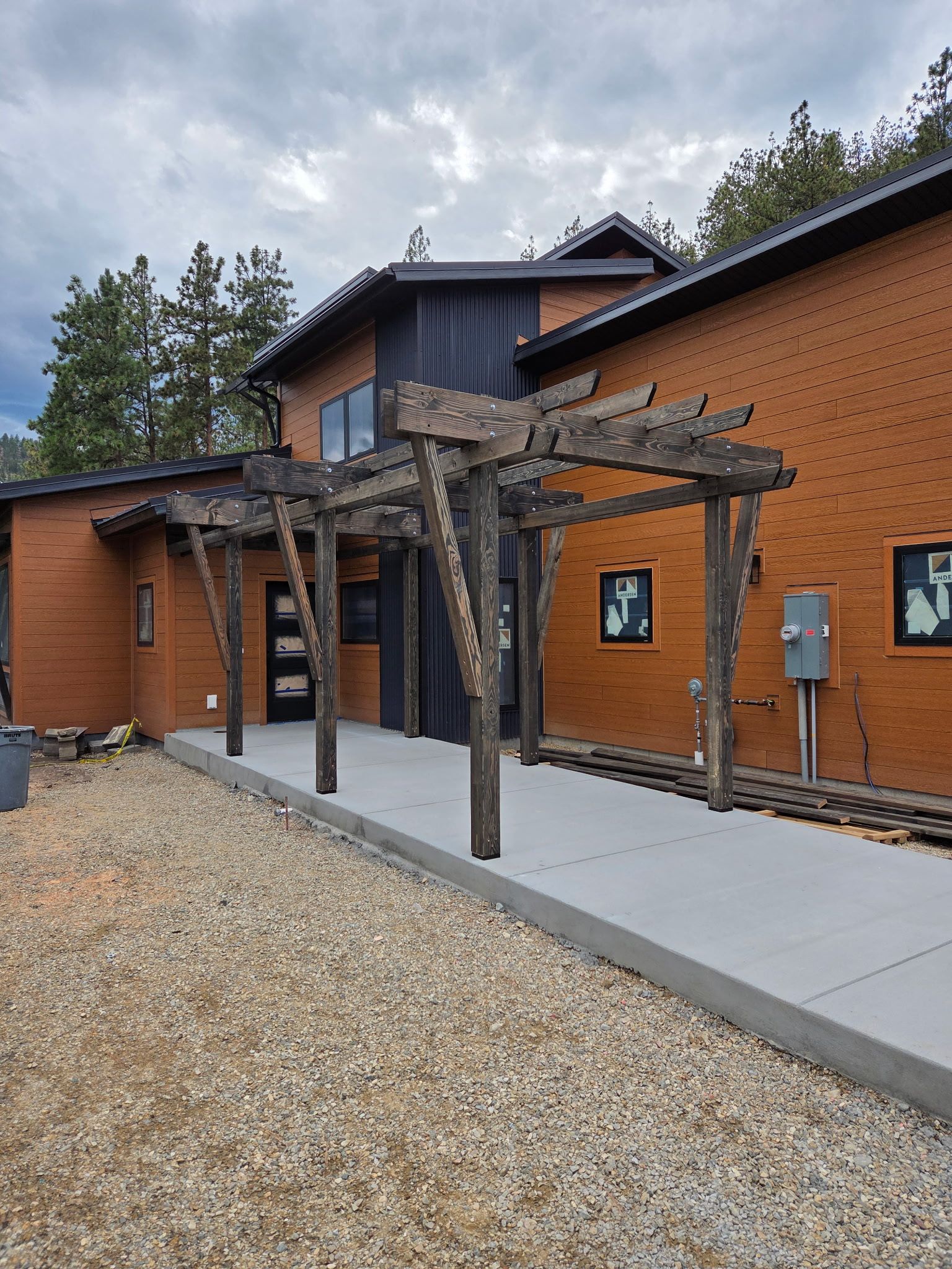 Brick building with wooden pergola over a concrete path; gray gravel foreground.