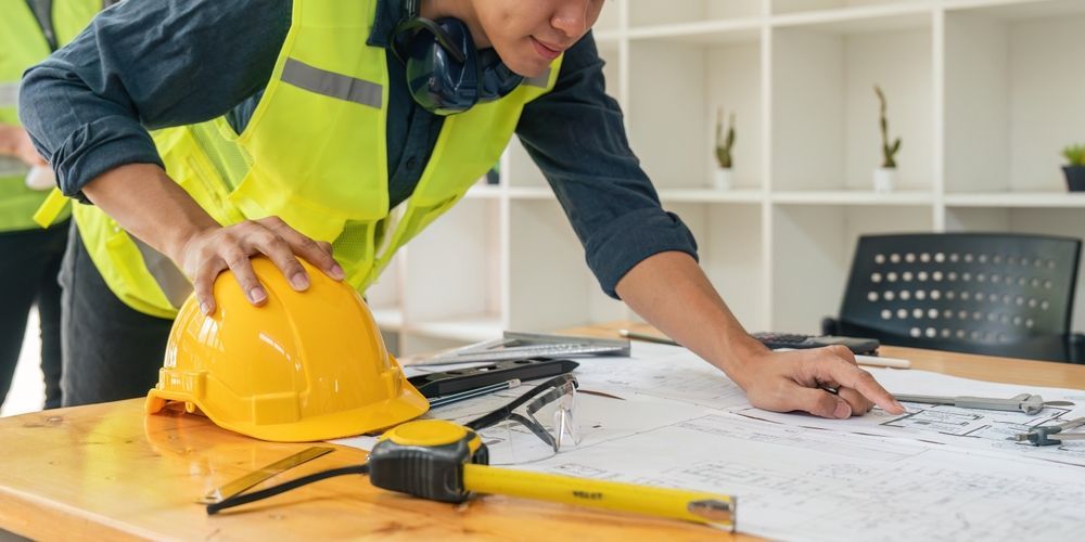 Person in safety vest pointing at blueprints on a desk with a yellow hard hat and tape measure in an office.