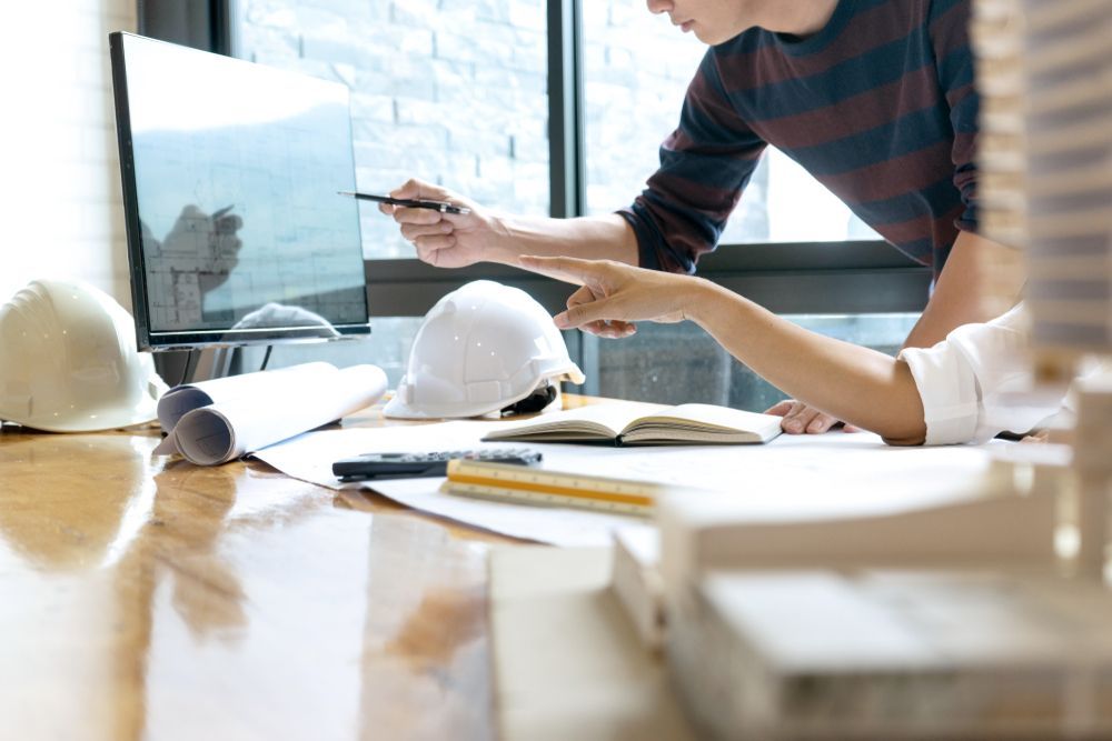 Two architects discussing a project, pointing at a computer screen. Hard hats, blueprints, and model on table.