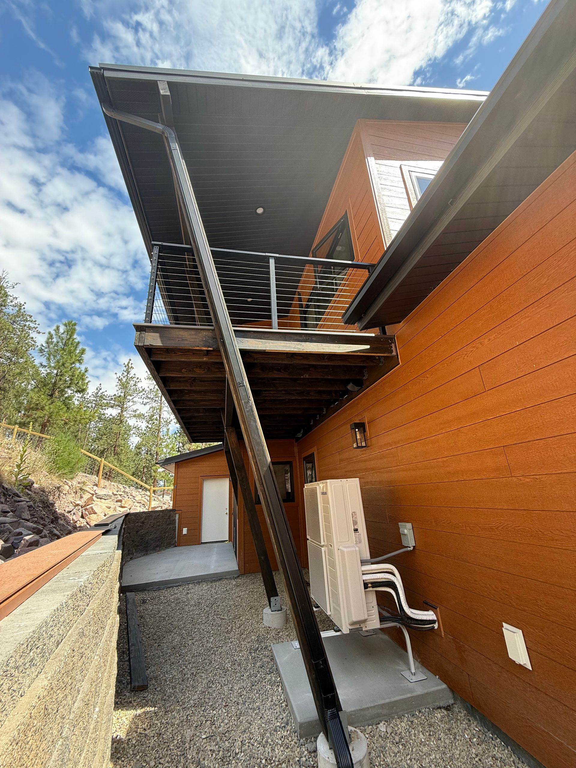 Exterior view of a two-story house with a dark wooden deck, a gravel path, and an air conditioning unit.