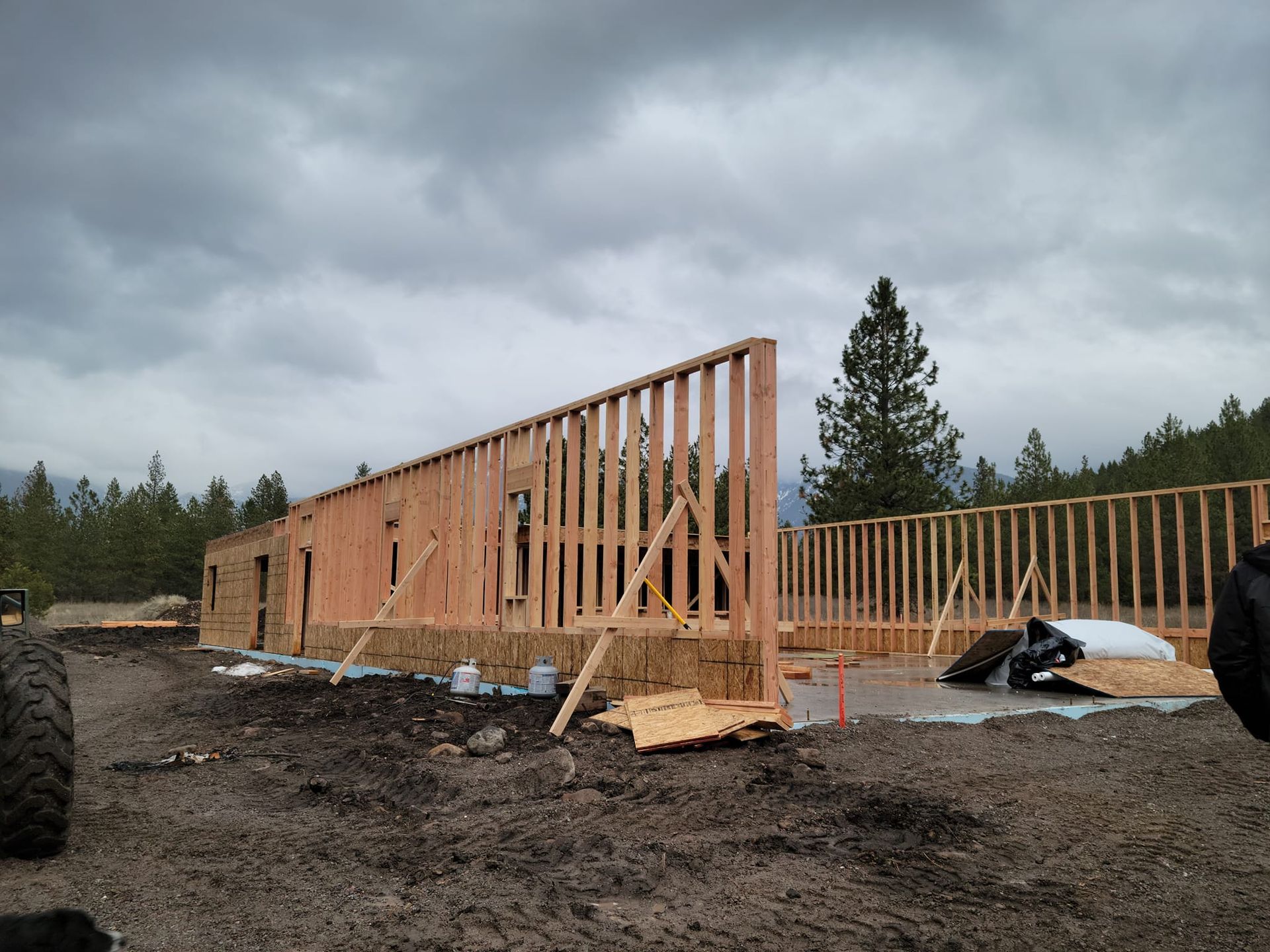 Wood frame of a building under construction on a muddy site, against a cloudy sky.
