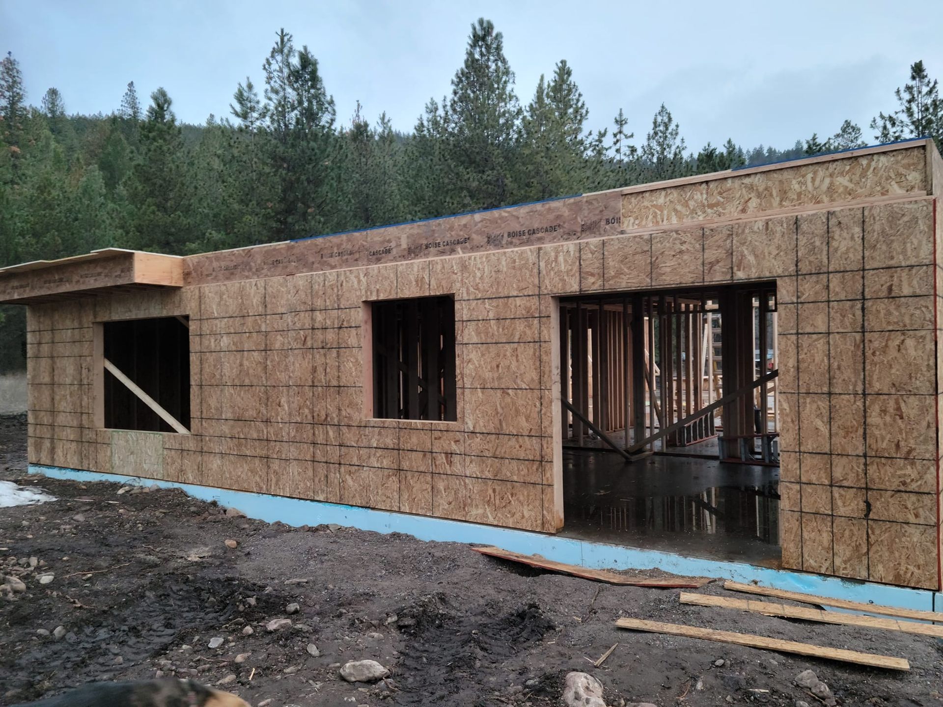 Construction site: building with OSB siding, windows, and a garage entrance, surrounded by dirt and trees.