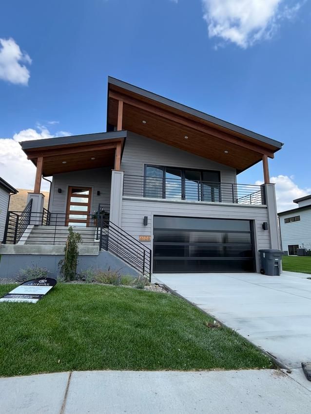 Modern gray house with wood accents, glass garage door, and black railings, on a sunny day.