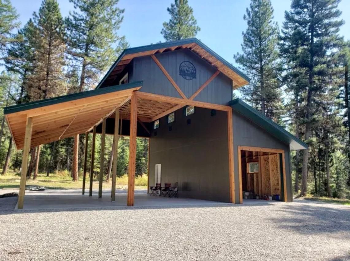 Gray barn-style building with a wooden carport, set in a forest with gravel driveway, blue sky.