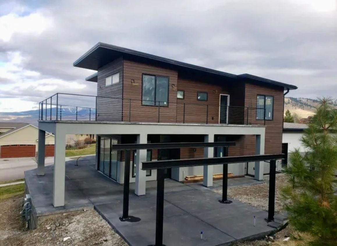 Modern two-story house with brown siding and a flat roof. Features a balcony with glass railings and a carport underneath.