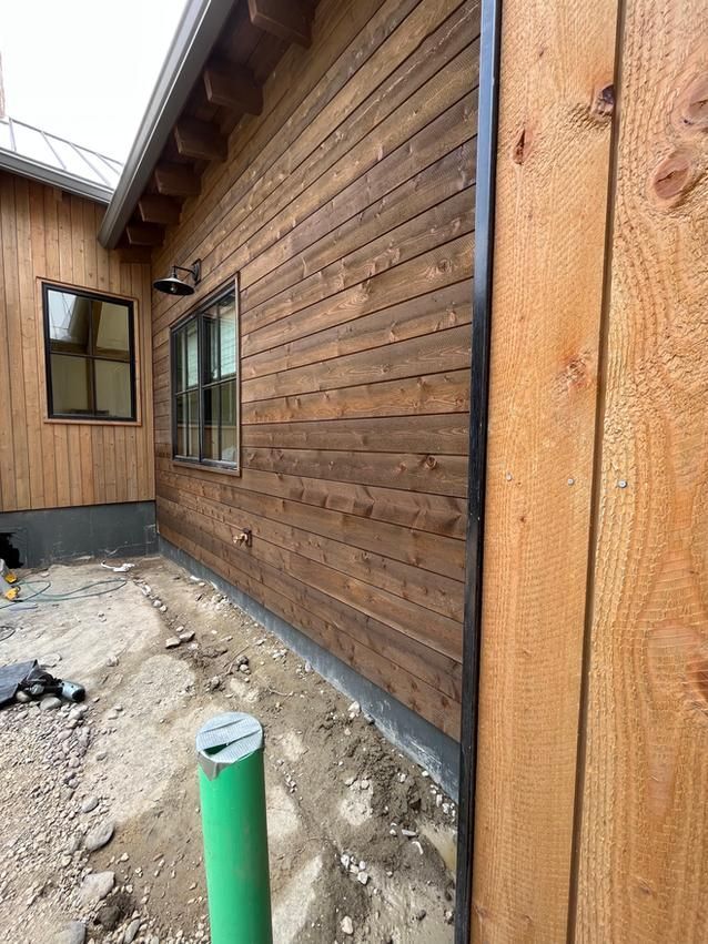 Exterior of a building with brown wood siding and a black trim; a green pipe is visible in the foreground.