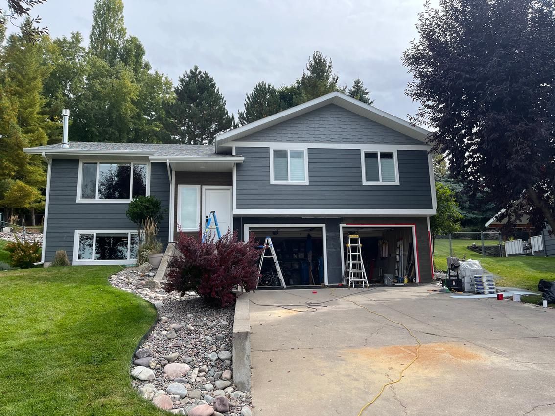 House exterior, gray siding, white trim, undergoing renovation. Garage doors open, ladder visible, cloudy day.