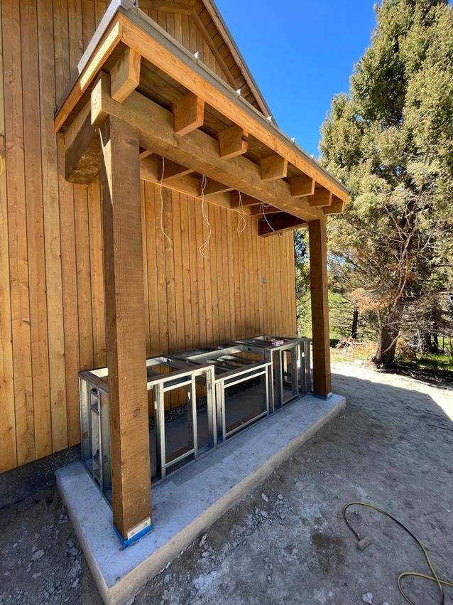 Outdoor kitchen with wooden pergola, stainless steel frames, and concrete base against a wood-sided building.