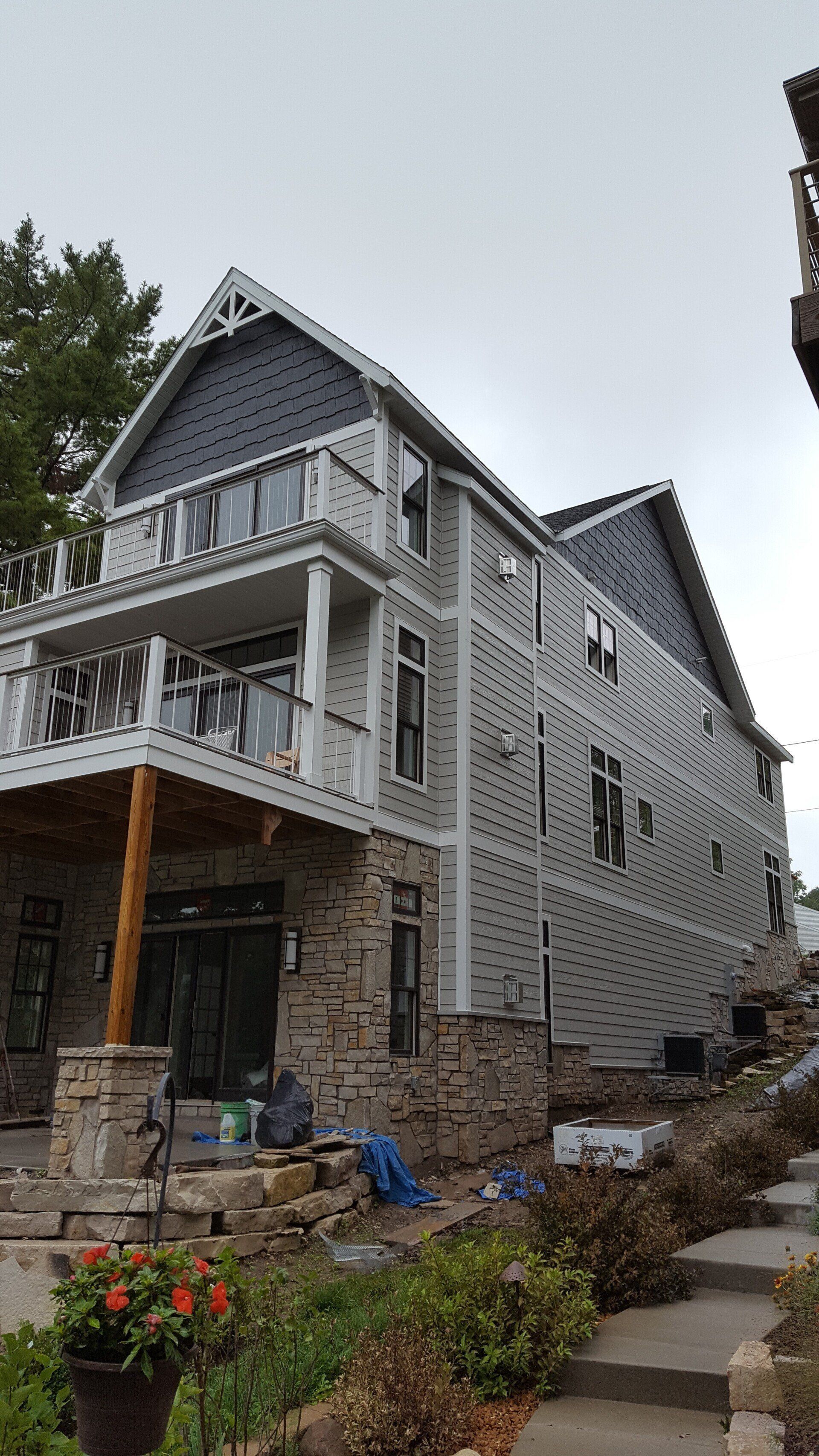 Three-story house under construction: gray siding, stone base, two-story balcony with white railings, blue tarp, cloudy sky.