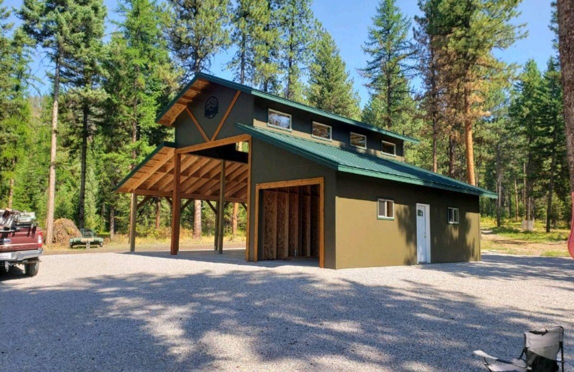 Green building with a carport, gravel driveway, and surrounding trees.