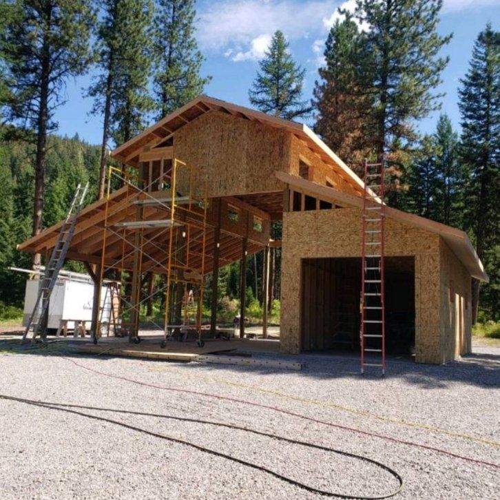 Partially constructed wooden barn with a garage, scaffolding, and ladders, set amongst trees.