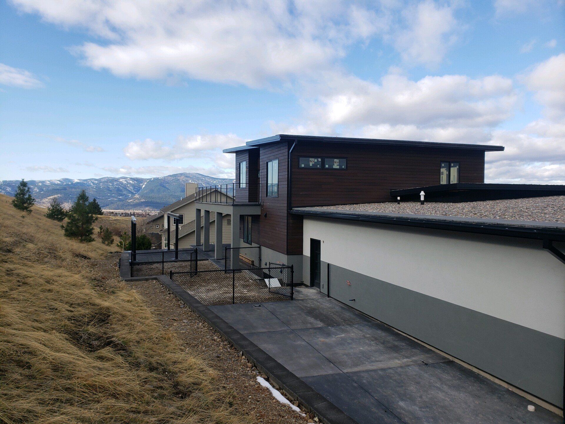 Modern house on a hillside with a driveway. Brown and gray exterior, blue sky, distant mountains.