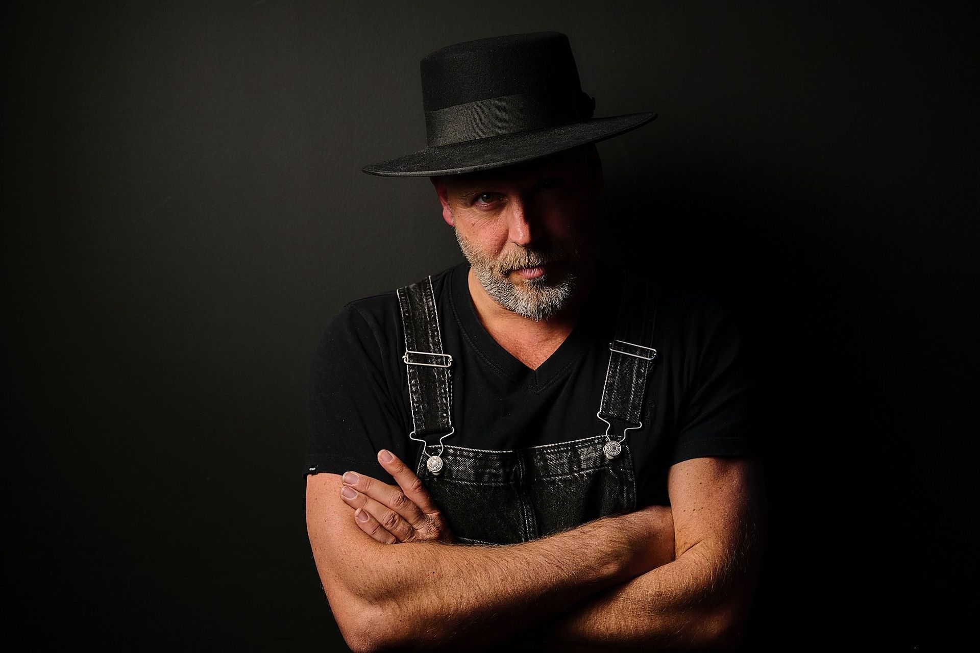 Man met zwarte hoed en shirt, armen over elkaar, tegen een donkere achtergrond.