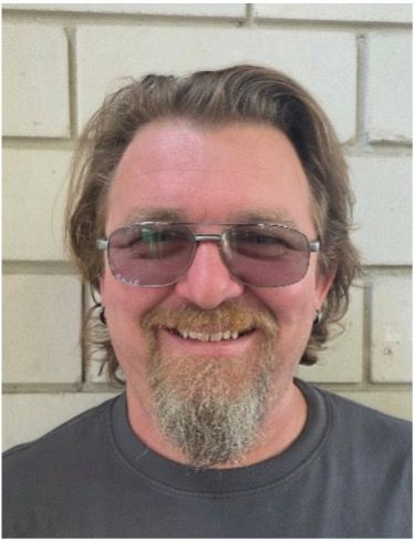 Smiling man with glasses and goatee standing against a light brick wall