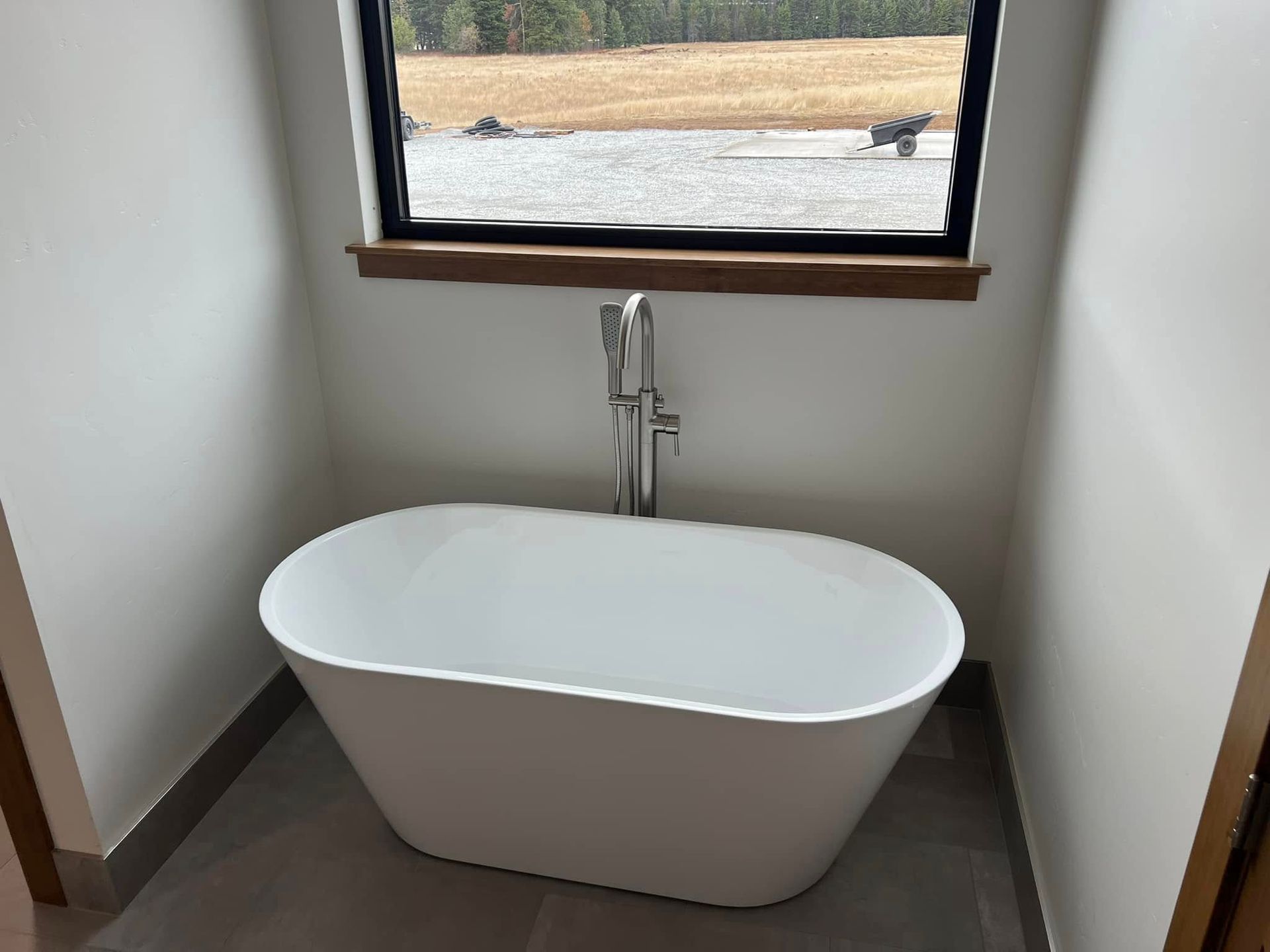 A modern white soaking tub sits beneath a window in a bathroom with light gray tile flooring.