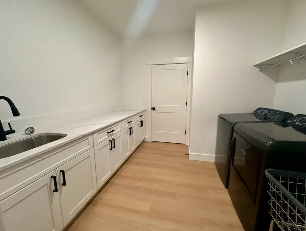 Laundry room with white cabinets, countertop, and walls. Black washer and dryer, and a door at the end. Light brown floor.