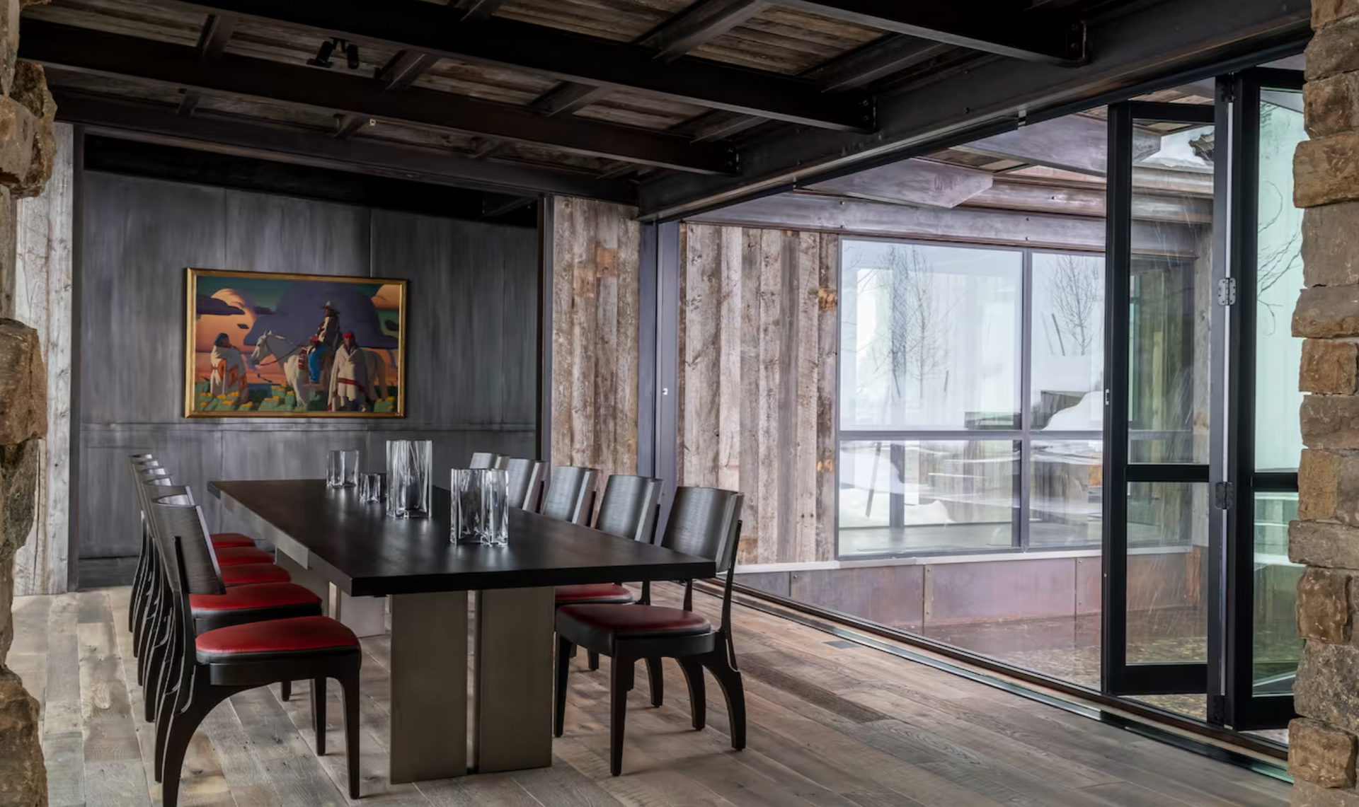 Dining room with long table, red chairs, art, wooden walls, and large window overlooking a wet outdoor space.