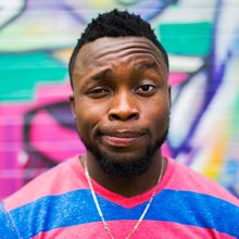 Man with short black hair and a beard, wearing a striped shirt, making a puzzled expression against a colorful graffiti wall.